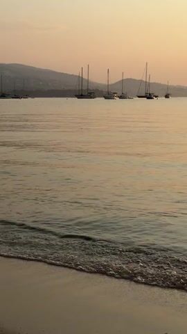 The video shows a tranquil scene at the beach during sunset. The calm sea gently laps at the sandy shore in the foreground, creating small waves. In the distance, a line of sailboats is anchored, their masts silhouetted against the warm, hazy sky. Mountains form a soft backdrop on the horizon. The overall ambiance is peaceful and serene, capturing the beauty of a coastal sunset.