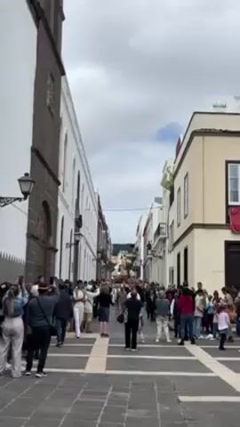 The video, filmed in a vertical orientation, shows a narrow cobblestone street lined with buildings, most of which have two stories. People of various ages and ethnicities are seen standing on the street, some holding phones or cameras. The camera is positioned at one end of the street looking down towards the other end. The sky is overcast with patches of blue visible. The architecture includes a tall, imposing building on the left with dark stonework, and lighter-colored buildings on the right. A rhythmic sound, possibly drumming, is audible.
