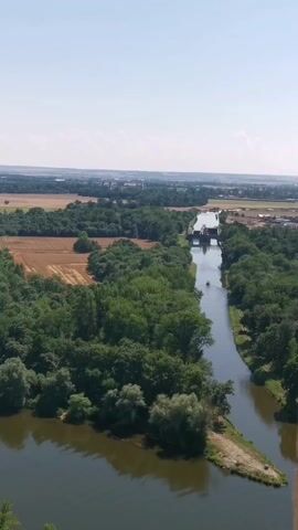 An aerial drone shot shows a wide river or canal with a narrow boat traveling on it. The boat is in the middle of the waterway, moving away from the camera. The waterway is flanked by lush green trees and some fields with dry vegetation. In the distance, there is a structure that appears to be a bridge or a lock, and beyond that, a hazy landscape with buildings and more fields. The sky is clear and blue, indicating a sunny day.