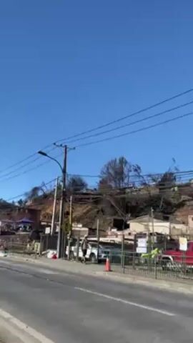The video shows a roadside view of a hillside neighborhood ravaged by fire. The sky is clear and bright blue. A two-lane road runs along the bottom of the frame. On the far side of the road, the lower parts of buildings and trees are visible. On the hill, scorched remains of buildings and trees are visible, and a few structures are still standing. Burned vegetation and debris cover the hillside. Utility poles and power lines are visible, as well as fencing and other roadside objects. People are walking on the side of the road. Several vehicles are parked or moving nearby.