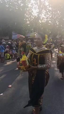 A group of women in traditional attire are walking in a parade on a sunny day. The woman in the foreground is wearing a black dress with gold embellishments and a yellow flower in her hair. She is carrying a basket with food and drinks. The other women are also dressed in similar outfits and carrying baskets. In the background, there are spectators and vendors. The overall atmosphere is festive and celebratory.
