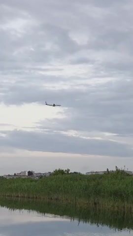 A plane flies across a cloudy sky above a body of water reflecting the sky and a line of tall green grass. Structures and trees are visible in the distance beyond the grass. The plane is white with a dark tail and what appears to be a red stripe on its tail.