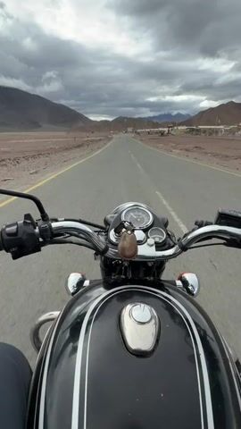 This is a first-person perspective video of someone riding a black motorcycle with silver pinstripes down a paved road. The rider's hands are visible on the handlebars, which are chrome and feature a speedometer and a small brown keychain. The motorcycle's fuel tank is in the foreground. The road stretches into the distance and is bordered by dry, barren land with mountains in the background. The sky is cloudy and overcast. There are a few signs and buildings visible along the side of the road. The video appears to be shot in a remote, mountainous region.