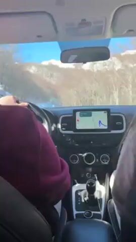 The video shows a view from inside a car, presumably the driver and passenger seats, as it drives on a snowy road. The driver, wearing a burgundy sweatshirt, is holding the steering wheel. Next to them, a passenger with long hair is seen. A GPS navigation screen is visible on the dashboard, displaying a route. The outside environment appears to be a mountainous area with trees covered in snow. The sky is clear and blue.