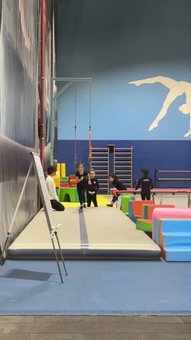 The scene captures a gymnastics training facility indoors. A white whiteboard on a tripod stands in the foreground, slightly obscuring the view. To its right, a young woman kneels, possibly instructing or observing. Three young girls in matching black outfits are on a long, gray mat with a dark blue stripe. One girl performs a handstand, the second stands beside her, and the third is further behind on the mat. In the background, there are colorful foam blocks, a wall ladder, a trampoline, and a blue wall with a white silhouette of a gymnast. Another girl is exercising on the trampoline. The flooring is dark blue. The walls have a patterned texture, and the lighting seems artificial.