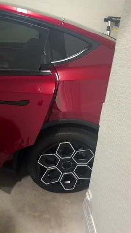 The video shows the interior and exterior of a red electric car, likely in a garage. The car has a sleek design with black accents on the doors and wheels. The interior features white seats, a large touchscreen display, and ambient red lighting. The camera pans across the car's exterior, highlighting the distinctive wheels and the open driver's side door, before moving inside to show the dashboard and steering wheel.