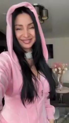 A woman with long black hair and light skin smiles at the camera while wearing a pink hooded jacket and a sparkly choker. She is indoors in a room that appears to be a living or dining area. Behind her are flowers in a vase on a table, a dining table with chairs, a chandelier, and a television mounted on the wall. The woman smiles at the camera and the view is mostly from the chest up.
