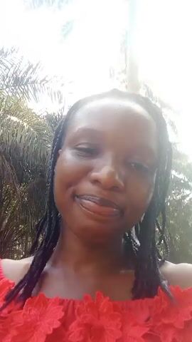 The video features a woman with dark skin and braided hair wearing a red off-the-shoulder top with flower-like embellishments. She is outside with trees and bright sunlight in the background. At one point, a child's face briefly appears behind her. The woman smiles at the camera.