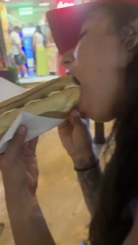 The video shows a close-up of a person eating a hot dog. The person is wearing a red cap and has long dark hair with tattoos on their arms. They are holding the hot dog with a white napkin and taking a bite. The hot dog is in a bun with ketchup and mayonnaise. In the background, there are blurry figures of people standing outside a food stall or shop. The environment appears to be an outdoor setting with warm lighting.