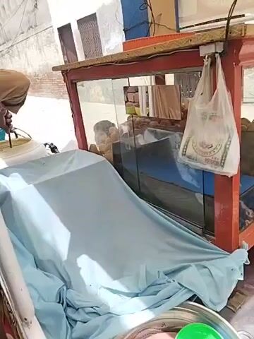The scene is a close-up view of a food cart selling gol gappe, an Indian snack. A red framed glass display case contains fried, round snacks inside.  A blue cloth drapes over part of the cart. In the foreground is a silver pot containing pink and green bowls and plates.  To the right, two hands are preparing gol gappe on a pink plate. The snack is being filled.  The environment appears to be outdoors, possibly a street.