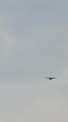 A long-shot video shows an airplane flying across a grey, overcast sky. The airplane is visible on the right side of the frame, with its wings spread and its body appearing dark against the sky. The airplane is flying from right to left across the screen. The sky is the dominant feature of the video, with no other objects or people visible.