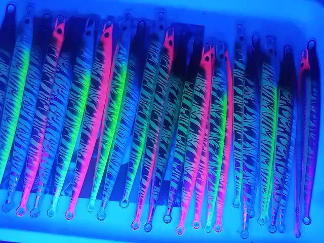Here is a description of the video:

The video shows a rectangular tray filled with fishing lures under a bright blue light. The lures are long and slender, with a tear-drop shape to their ends.  They appear to be arranged in rows, and some are various colors like green, pink and blue.  Each lure has a black tiger stripe pattern along its length. Some lures are lit up with a bright green glow, especially when the light shifts.  All the lures appear to have small loops at the top for attaching fishing lines.  The overall impression is a display of vibrant, eye-catching fishing equipment.