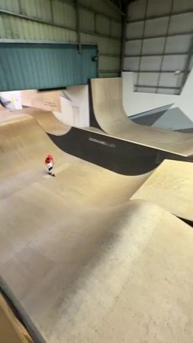 Here's a concise description of the video:

The video shows a young person wearing a red t-shirt, white pants, and an orange helmet riding a scooter in an indoor skatepark. The person rides up a ramp, jumps into the air, and lands smoothly. They then ride along a curved wall, ending at the bottom. The skatepark has various ramps and curves, and logos are visible on the walls.