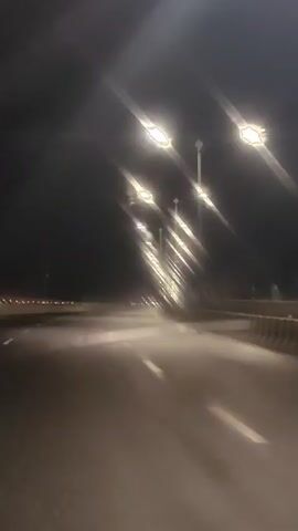 The video shows a blurry night view of a road from the perspective of a moving vehicle. Streetlights line the road, casting a bright glow that creates streaks of light. The road itself is dimly lit, with white lines indicating lanes. There are some faint lights visible in the distance, suggesting it might be a highway or a road in a populated area. The overall atmosphere is dark and somewhat ethereal due to the lighting and blurriness.