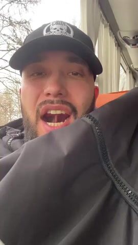 A man wearing a black baseball cap, a black jacket, and a gray beard and mustache opens his mouth very wide, revealing his teeth, tongue, and the inside of his mouth. He is sitting inside a bus with an orange seat behind him and a window to his right. Bare trees are visible through the window.
