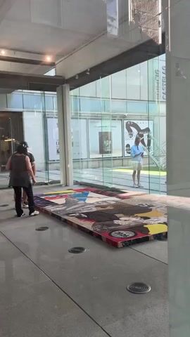 The video shows an interior of a modern building, likely an art gallery or museum, with a polished concrete floor and large glass windows. Two individuals are walking away from the camera, and a third person is standing outside, looking into the building. In the foreground, there is a large, colorful rug or tapestry laid out on the floor, featuring a collage of images and abstract designs. The rug is a focal point of the scene. The lighting appears to be natural, coming from the large windows.