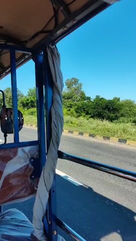 The video is filmed from inside a vehicle, looking out the front. The vehicle appears to be a three-wheeled auto-rickshaw or similar open-air transport. The camera is positioned behind the driver, who is wearing a grey t-shirt and has short dark hair. The driver is facing forward, looking at the road ahead. The interior of the vehicle has blue metal bars and a clear plastic shield with the words "MAX POWER" printed on it in large red letters. There is a small electric fan mounted on the left side of the vehicle's interior. Outside the vehicle, a paved road stretches into the distance, flanked by grassy verges and trees. The sky is blue and clear, indicating it is daytime. The video captures the movement of the vehicle as it travels down the road.
