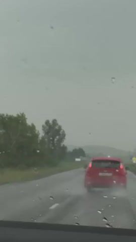 The video shows a view from inside a car driving on a wet road in the rain. A red car is visible ahead on the road. Trees and a cloudy sky are visible in the background. The windshield wipers can be seen moving across the glass, indicating the heavy rainfall.