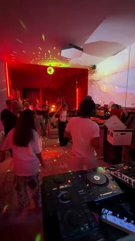 The video shows a group of people dancing in a dimly lit room. The room is decorated with red and green lights, and a disco ball is hanging from the ceiling, casting colorful reflections on the walls. In the foreground, a DJ setup is visible, with turntables and a mixer. Several people are standing and dancing in the room, some of whom are wearing casual clothing. The overall atmosphere appears to be a lively party.
