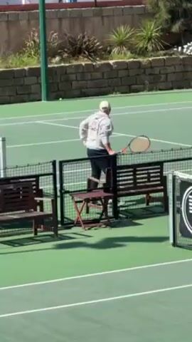 A man stands next to tennis court benches, holding a tennis racket. He is wearing a gray sweatshirt that says "COACH" on the back, along with dark pants and a light-colored hat. He walks towards the camera, then appears to hit a tennis ball. The camera points down at the tennis court, then rotates to show a closeup of dark, slatted wood.