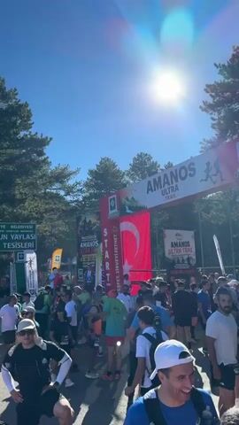 This video captures a vibrant outdoor event, likely a race start, with a large crowd gathered under a clear blue sky. Numerous participants, dressed in athletic wear and carrying backpacks, mill about in anticipation. Several large banners and flags are visible, including Turkish flags and those displaying event branding, such as "AMANOS ULTRA." A prominent archway with "START" written on it suggests the beginning of a course. Behind the crowd, tents and structures indicate a well-organized event setup. Tall trees form a natural backdrop, and the ground appears to be paved or a dirt path. The atmosphere is energetic and communal, with people interacting and preparing for an athletic endeavor.