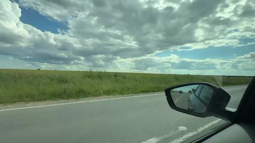 The video captures a view from inside a car, looking out the passenger side window and into the side-view mirror. The car is traveling on a paved road that runs alongside a vast, green field of crops, possibly wheat or corn, stretching to the horizon. The sky above is filled with dramatic, white, fluffy clouds against a bright blue backdrop. The side-view mirror reflects the road ahead, showing another car in the distance and the same expanse of fields and sky. The overall impression is a scenic drive through a rural landscape on a cloudy but bright day.
