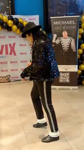 A man dressed as Michael Jackson performs a dance routine in front of a Muvix banner and a Michael Jackson poster. He wears a black sequined jacket, a white shirt, black pants with a silver stripe, a black fedora, and white socks with black shoes. He is performing on a tiled floor, with a balloon arch made of gold and black balloons behind him. A speaker is visible to his left, and a few people are in the background, including a child sitting on the floor. The Muvix banner has the Muvix logo repeated multiple times. The Michael Jackson poster displays an image of the singer and some text, including a QR code.