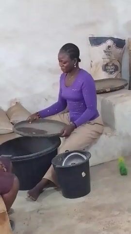 A woman wearing a purple long-sleeved shirt and khaki pants is sitting on a low concrete step indoors. She is barefoot and sifting something in a large, shallow metal pan. Underneath the pan is a large black bucket. Next to her is another black bucket with a lid. A woman wearing a dark shirt and purple pants is sitting next to her on a wooden stool. Bags of flour are stacked on the concrete step behind the woman doing the sifting. The setting appears to be a small, simple room.