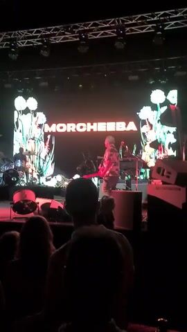 The video shows a dimly lit concert scene with a large screen in the background displaying the band's name, MORCHEEBA, in neon pink lettering, surrounded by animated floral graphics. A band member, wearing a colorful patterned shirt, is seen on stage in front of the screen, appearing to play an instrument. Another band member is visible behind a drum set on the left side of the stage, and other band members are positioned behind keyboards on the right. In the foreground, the backs of several audience members are visible, silhouetted against the stage lights. The overall atmosphere suggests a live musical performance in an indoor venue.