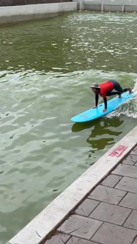 A person in a red long-sleeved shirt, black pants, and a patterned bucket hat is wakesurfing on a blue surfboard. They are positioned with their left foot forward, balancing on the moving board. A white wake is visible behind the board. The water is green and appears to be in a canal or man-made waterway, with a concrete wall visible on the right.