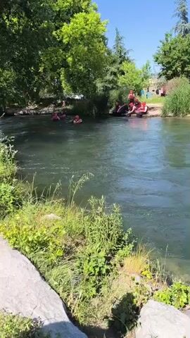 The scene depicts a riverbank setting on a sunny day. The river's water appears to be flowing with a noticeable current. On the opposite bank, a group of individuals, some wearing red inflatable personal flotation devices, are visible. Some appear to be in the water, while others are near a raft or boat on the river's edge. The riverbanks are adorned with green vegetation, including grasses, bushes, and trees. The sky is clear and blue, suggesting a bright, warm day. In the foreground, there is a large, gray rock and more green vegetation along the bank.