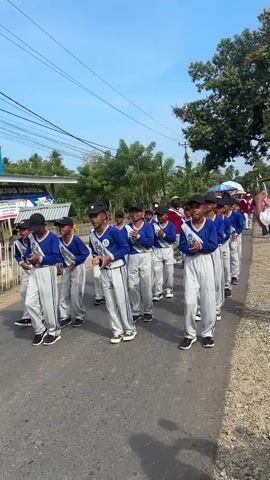 A group of young boys in matching blue shirts, grey pants with blue stripes, and black caps are marching in formation down a paved road. They are all wearing white sashes over their blue shirts. The boys are holding what appear to be small instruments or batons. The road is lined with a fence and some buildings on the left side, and trees and foliage on the right. The sky is clear and blue. The boys are moving from left to right in the frame.