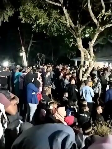 The video captures a large crowd of people gathered outdoors at night, illuminated by artificial lights, possibly from nearby buildings or streetlights. A large tree with prominent branches is visible in the foreground, casting shadows over the scene. The people are densely packed, with many facing towards a building in the background which appears to be a house or community center with a dark roof and illuminated windows. The attire of the individuals suggests a casual gathering, with a mix of genders and ages present. Some individuals are holding up phones, possibly recording or taking pictures. The atmosphere appears lively, with a sense of collective attention directed towards something happening in front of the building, though the specific activity is not discernible from the video. The ground is uneven and covered with dry grass or foliage.