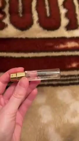 A hand holds a small, transparent perfume sample with a gold cap. The label reads "BYREDO" with "Rose of No Man's Land" written underneath. The background is a brown, tan, and red patterned textile, with what appears to be the word "ROCK" partially visible in a large, fuzzy font.