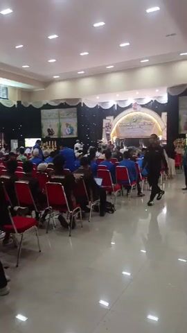 The video shows the interior of a large hall, likely a conference or event venue. The hall is filled with people seated in rows of red chairs. In the foreground, three young males are walking towards the camera. Two of them are wearing black jackets with a circular logo on the back depicting a red panda and text that appears to say "ONE DECADE YAKAM SYARIAH". The third male is wearing a brown jacket with a similar logo. The background features a stage area with a large banner displaying text and images, and a curtain backdrop. The lighting is bright, emanating from overhead fixtures. The overall atmosphere appears to be that of an organized gathering.