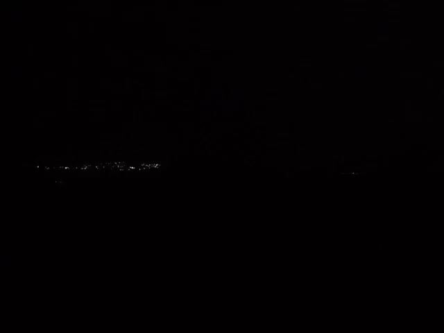 The video begins with a completely black screen. At the bottom of the frame, barely visible, is a line of faint lights, possibly from a distant city. Suddenly, a single streak of pink lightning flashes brightly in the center of the screen. The lightning disappears quickly, and the scene returns to the initial black screen with the dim city lights.