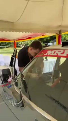 A man is working inside the cockpit of a rally car under a tent. Another man is seen standing on top of the car. The car is white with red and green stripes and says "MRF TYRES" in large red lettering across the top of the windshield. The car also has emergency contact information on the driver's side. The tent is white with yellow and red accents. Trees and green grass are visible in the background.