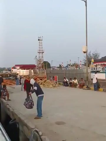 A wide shot shows a busy port scene. In the foreground on the left, the white hull of a large boat is visible. A concrete pier runs along the bottom of the frame. On the pier, a group of people are gathered, some sitting and some standing. A woman in a blue hoodie and jeans is standing near the edge of the pier, holding a patterned tote bag. Further back, there are more people, some appearing to be workers, moving around. Behind them, construction materials, including wooden planks, are stacked. A tall tower with some equipment on top stands in the middle ground. To the right, several buildings are visible, one with a traditional-looking roof. A large construction crane is positioned behind the buildings. More people are walking and standing in this area, and several motorcycles are parked. The sky is overcast and the lighting suggests it is either early morning or late afternoon.