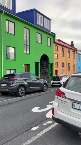 The video shows a street view in what appears to be a European city, judging by the architecture and car license plates. The buildings are brightly colored, with one building being a vibrant green, another orange, and another blue. A dark grey Hyundai Kona is parked in front of the green building. A white Toyota Aygo is parked nearby, and a white Subaru Outback is in the foreground. The green building features an archway with a mural inside, depicting a cartoonish monster with a large, toothy grin. The street has a white arrow painted on it. The sky is overcast.