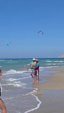 The video opens with a wide shot of a beach on a sunny day. The water is a clear turquoise, and there are waves breaking on the shore. In the foreground, a woman in a black swimsuit and white cover-up walks towards the water. Further down the beach, two men are standing in the shallow water, appearing to be instructing or assisting someone in kitesurfing. One man, wearing a yellow hat, is holding a kite control bar. In the distance, several kites are visible in the sky, indicating kitesurfing activity. The camera then pans to the ocean, showing the vast expanse of blue sky and water, with kitesurfers appearing as small colorful dots. It then focuses on a purple kite floating in the water near the shore. The video concludes with a woman in a yellow dress standing by the water, holding up a phone to take a picture. The environment is a sunny beach with a clear blue sky, calm ocean, and some hills in the background.