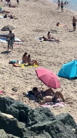 The video shows a crowded beach scene. In the foreground, a pink umbrella shades two people lying on towels. To their right, a blue beach tent is set up with one person inside. Further up the beach, several other individuals and small groups are sunbathing on towels. In the background, the ocean is visible with people swimming and a few individuals standing near the shoreline. The sand is light brown, and there are some rocks in the immediate foreground. The sky is bright and appears to be sunny.
