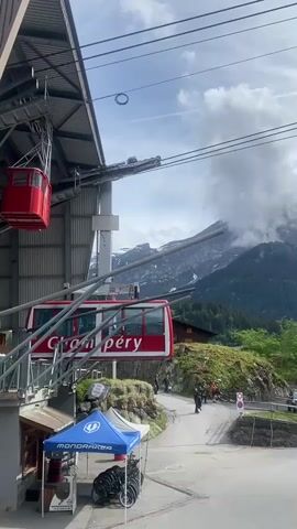 A red gondola car is prominently featured at a station, with "Cham péry" written on its side. Another red gondola car is partially visible higher up near the station structure. In the background, majestic mountains rise, their peaks partly obscured by clouds. Lush green trees cover the lower slopes of the mountains. Below the gondola station, a paved path or road winds, with several people walking along it. A blue tent with "Mondraker" written on it is set up near the ground level, with what appears to be bicycles displayed or stored nearby. The sky is partly cloudy, suggesting a daytime scene. The overall setting is a mountain village, likely a ski or bike resort.