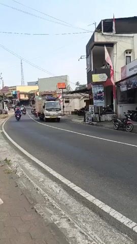 A short video captures a street scene during daylight. In the foreground, a gray concrete sidewalk with a white stripe runs along the left edge of the frame. The main focus is a paved road with clear lane markings. Several motorcycles are seen on the road. Closest to the camera, a group of motorcycles is traveling away from the viewer. Further down the road, a white truck is traveling in the same direction, carrying a large load of stacked lumber. The truck appears to be overtaking the motorcycles. On the right side of the road, a building with multiple stories is visible, featuring a storefront with signage that includes the words "KOMPO MOTOR - SAPURAN". Several motorcycles are parked in front of this building. Two red and white flags are displayed on poles attached to the building. The sky is clear and bright, suggesting a sunny day. The overall impression is of a typical street scene in a populated area, likely a town or city, with ongoing traffic and commercial activity.