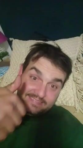 A man with dark, messy hair and a beard smiles and gives a thumbs up. He is wearing a green shirt and is in front of a white, textured pillow. He then runs his hand through his hair.
