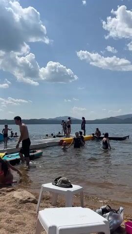 The video shows a sunny day at a lake with many people enjoying various water activities. A group of people are gathered on a wooden dock extending into the water. Some are standing and talking, while others are swimming or playing in the water. Two children are sitting on the sandy shore in the foreground, one with an orange inflatable ring. A man in black shorts is standing near the edge of the dock, looking towards the water. A blue slide is visible on the left side of the frame, with a few people near it. The lake is calm and stretches out towards distant, hazy hills under a bright blue sky with fluffy white clouds. In the foreground, two white plastic chairs are placed on the sand, one with a dark bag on it.