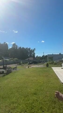 This media file captures an outdoor scene on a bright, sunny day in Khvoynoye, Russia. The sun is the dominant subject, appearing as an intense, radiant orb that creates lens flare and washes out parts of the image. The sky is a clear, vibrant blue, with a few wispy clouds present.

The setting appears to be a yard or outdoor area, likely residential, with a well-maintained green lawn. In the foreground, a person's foot is visible at the bottom left, suggesting they are relaxed and enjoying the outdoors.

Scattered around the yard are various objects. There's a metal grill or barbecue setup, accompanied by what appear to be two chairs, implying a space for outdoor cooking and relaxation. A fence lines the edge of the property, separating it from a wooded area or a row of trees that forms the backdrop. Beyond the trees, glimpses of buildings and a dark-colored car suggest a rural or semi-rural environment. The overall atmosphere is peaceful and serene, indicative of a warm, pleasant day. The time of day is clearly midday, given the high position of the sun and its intense brightness. No text is visible in the media file.