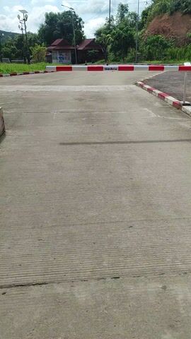 This video shows a barrier pole with red and white stripes, likely a gate or bollard, in Xayden, Laos. The pole has the word "Dashou" printed vertically in black text. The scene is outdoors on a paved surface, possibly a road or parking area. To the left of the pole, there's a grassy verge with decorative lampposts and some low-lying buildings with red roofs visible in the distance. Lush green trees and vegetation are abundant, with a hill or mountain in the background. The sky is partly cloudy, suggesting it could be late morning or early afternoon. There are no people visible in the video. The overall atmosphere appears peaceful and rural.