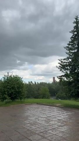 The video presents a stationary shot of a paved square. The square is made up of rectangular gray tiles. It appears to be wet, suggesting it has recently rained. Beyond the square, there's a grassy area with trees of varying sizes. In the distance, a brick tower is visible. A road curves through the trees. The sky is mostly covered with gray clouds, with a small patch of blue visible. There are no people present in the shot.