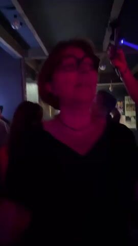 The video shows a woman wearing glasses and a black top dancing in a dimly lit venue with a wooden ceiling and exposed beams.  Other people are moving in the background, and there are colored lights flashing.