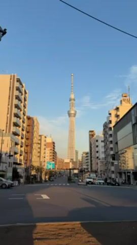 The video shows a street view in Japan, featuring the Tokyo Skytree in the distance. The street is bordered by buildings of varying heights and architectural styles. Several cars pass by on the road. The sky is a clear blue.